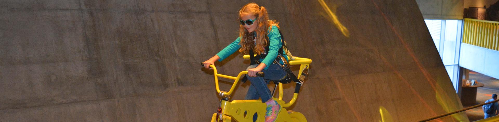 A blind girl rides a bike on a track that floats above a museum floor. 